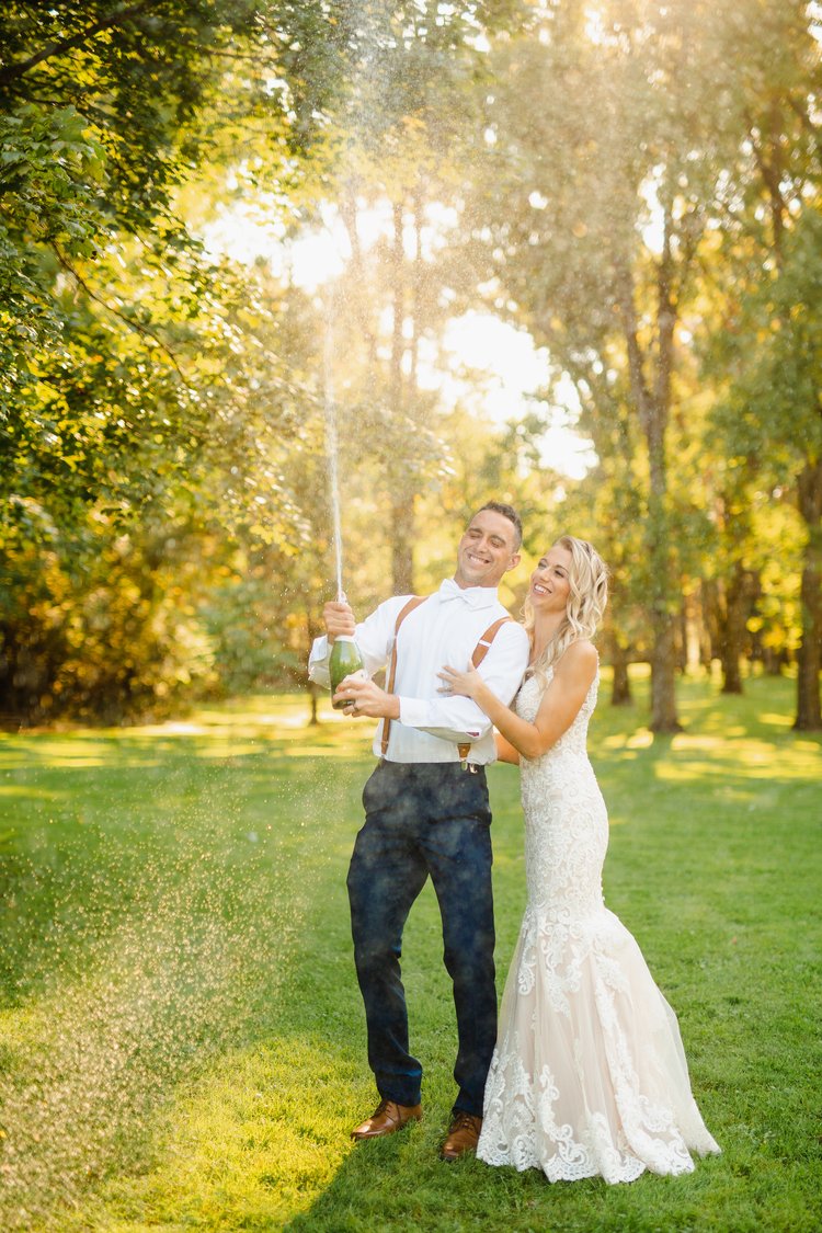 A newly married couple spraying a bottle of champagne in a forest.